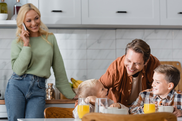 blurred woman talking on mobile phone near happy husband standing near sons having breakfast in kitchen - Foto, afbeelding