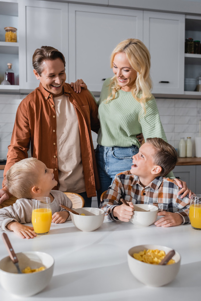 ευτυχισμένο ζευγάρι κοντά γιους τρώγοντας νιφάδες καλαμποκιού και πίνοντας χυμό πορτοκαλιού κατά τη διάρκεια του πρωινού - Φωτογραφία, εικόνα