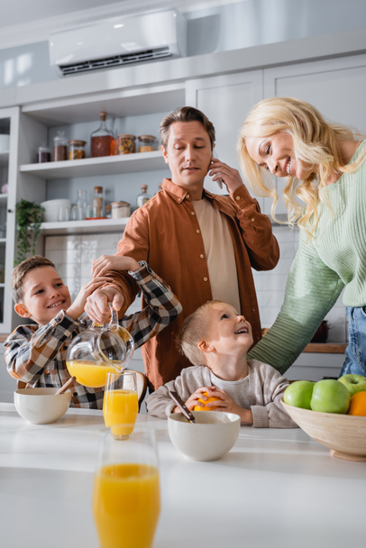 man pouring orange juice while talking on smartphone during breakfast with family - Foto, Imagem