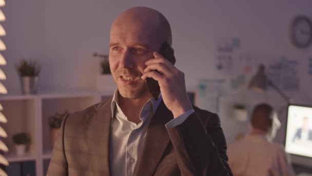 Medium close-up with slowmo of serious mid-adult businessman making phone call standing in dark office at night. His African-American business partner video conferencing in background - Footage, Video