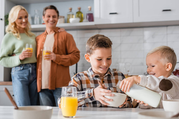 chłopiec wylewając mleko do miski podczas śniadania z bratem w pobliżu rozmazanych rodziców - Zdjęcie, obraz