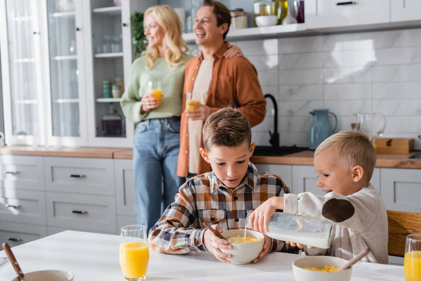 boy pouring milk into corn flakes while breakfasting with brother near blurred family - Foto, imagen
