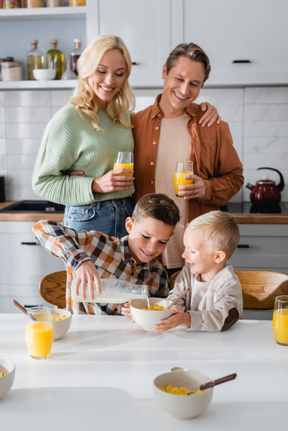 happy couple looking at son pouring milk during breakfast with brother - Foto, imagen