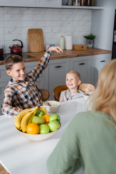 smiling boy pointing with finger at brother during breakfast with cereals and fresh fruits ner blurred mom - 写真・画像