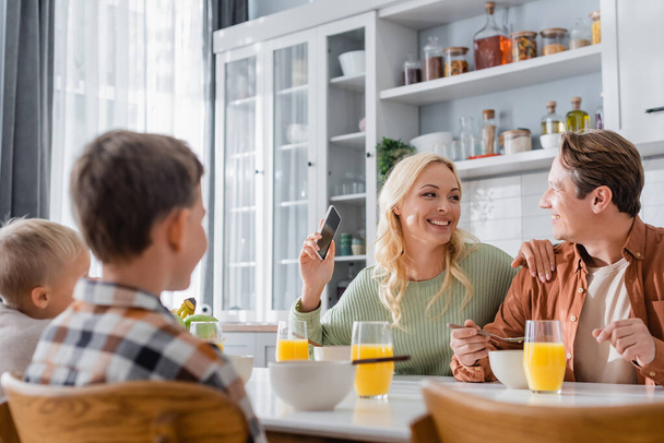 joyful woman with mobile phone looking at husband during breakfast with blurred kids - Foto, Bild