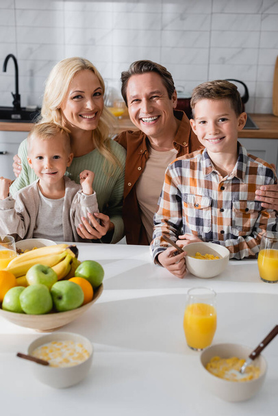 joyful couple with sons looking at camera near fresh fruits and orange juice in kitchen - Foto, Imagen