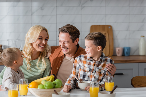 familia feliz riendo en la cocina cerca de vasos con jugo de naranja y tazón con frutas frescas - Foto, Imagen
