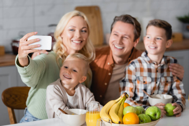 joyful woman taking selfie on cellphone with family near fresh fruits during breakfast - 写真・画像