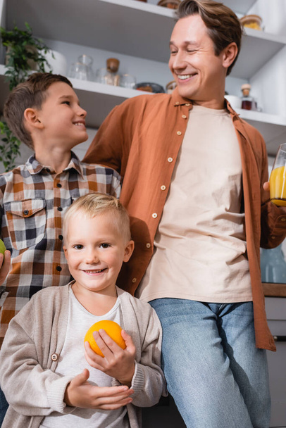 happy boy holding orange near blurred dad and brother in kitchen - Foto, afbeelding