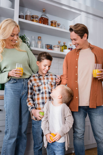 χαμογελαστό ζευγάρι με χυμό πορτοκάλι χαμογελώντας κοντά γιους κρατώντας φρούτα στην κουζίνα - Φωτογραφία, εικόνα