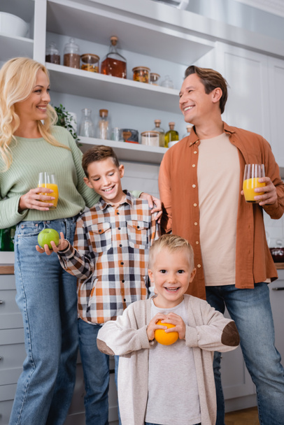 parents with orange juice and boys with oranges smiling in kitchen - Fotó, kép