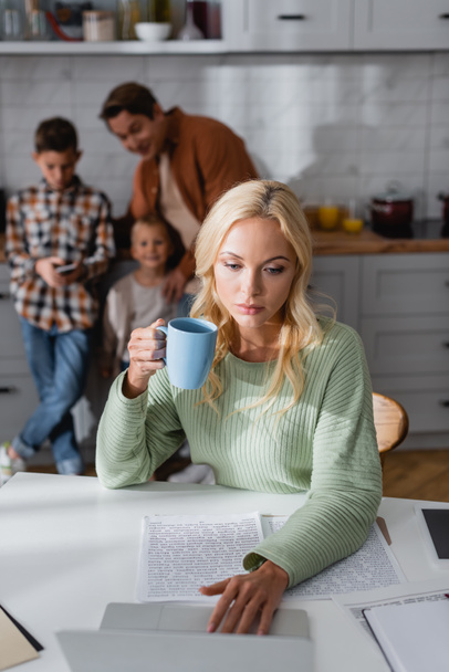 woman with cup of tea working near documents and laptop near blurred family in kitchen - Фото, зображення