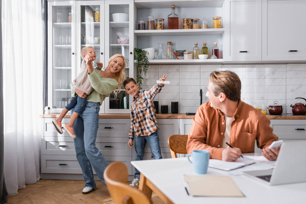 cheerful woman having fun with sons near blurred husband working in kitchen - Fotoğraf, Görsel