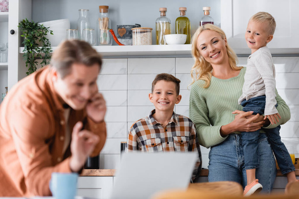 burred man showing okay gesture while talking on smartphone near laptop and family in kitchen - Photo, Image
