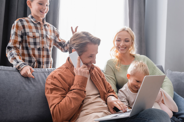 cheerful boy showing bull horn gesture near father working on laptop near family on couch - Photo, Image