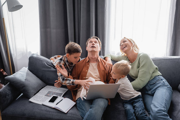 busy and tired man with laptop trying to work near cheerful family playing fun on sofa - Photo, Image