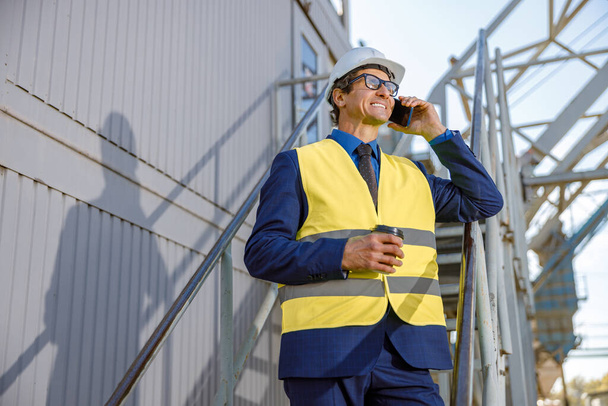 工場で屋外で携帯電話で話す陽気な男性エンジニア - 写真・画像