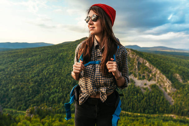 Portrait of a traveler in tourist gear. A young tourist with a backpack admires the sunset from the top of the mountain. Tourist woman with a backpack on a mountain background - Фото, изображение