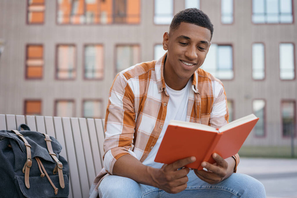 Porträt eines jungen, gut aussehenden Mannes, der Buch liest und sich im Park entspannen kann. Lächelnd selbstbewusster afroamerikanischer Student, der studiert, Sprache lernt und auf dem Universitätscampus sitzt, Ausbildungskonzept - Foto, Bild