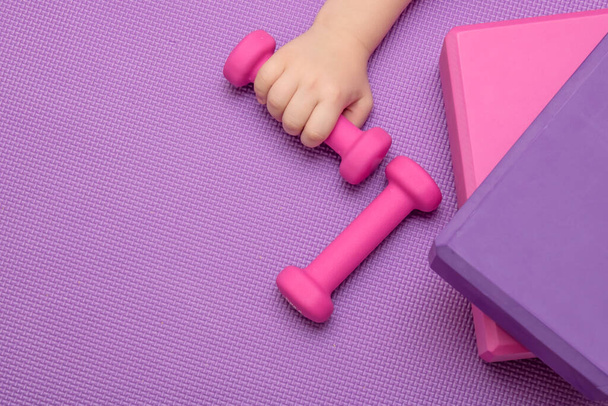 Sports equipment for fitness and yoga - dumbbell, blocks, one of which is held by a childs hand on a lavender-colored sports mat. Place for your text. - Photo, Image