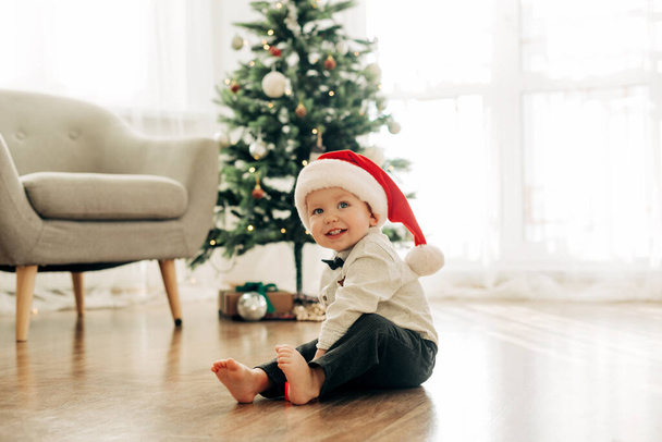 A laughing boy in a Santa Claus hat sits on the floor against the background of a Christmas tree - 写真・画像
