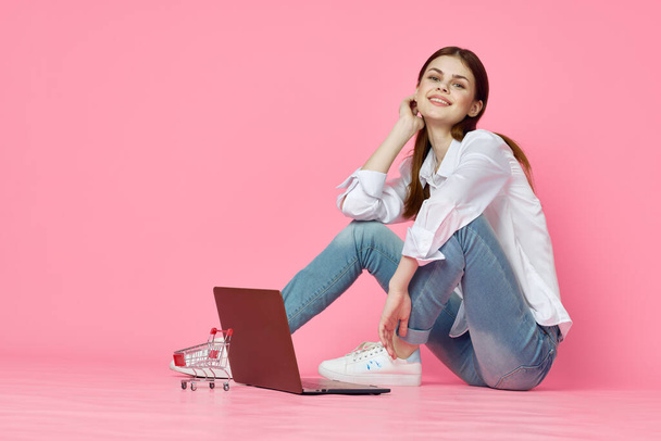 cheerful woman with laptop sitting on the floor on pink background internet technology - Фото, изображение