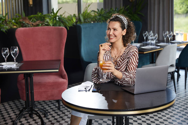 Krásná mladá kudrnatá žena sedí v kavárně a pije čerstvý pomerančový džus a čeká na svého přítele - Fotografie, Obrázek