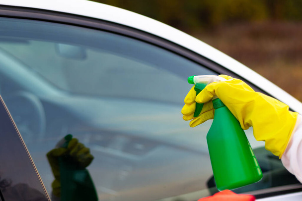 Рука в жёлтой резиновой перчатке брызгает водой из баллончика на окно автомобиля в тёплый осенний день. Мокрая уборка. Выборочный фокус. Крупный план - Фото, изображение