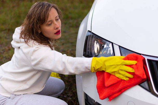 Ładna młoda kobieta w białej bluzie z kapturem i żółtych gumowych rękawiczkach wyciera reflektory samochodu jasnoczerwoną szmatą w ciepły jesienny dzień. Wybiórcze skupienie. Zbliżenie - Zdjęcie, obraz
