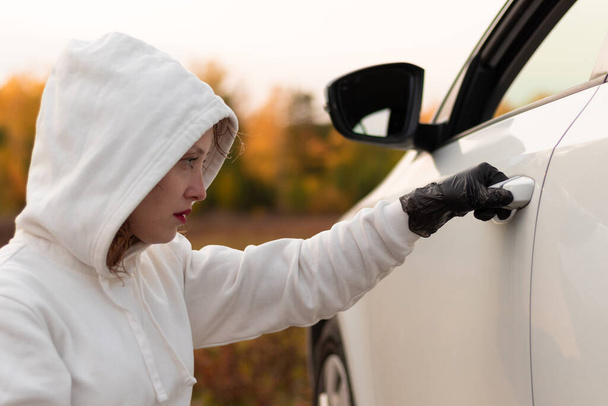 Kaunis nuori nainen valkoisessa puserossa, jossa on huppu ja mustat käsineet, avaa auton oven varastaakseen sen. Valikoiva keskittyminen. Lähemmäs. Muotokuva - Valokuva, kuva