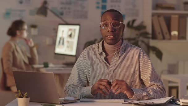 Média POV tiro de jovem afro-americano em óculos olhando para câmera discutindo documentos de negócios sentados na mesa no escritório escuro, enquanto sua secretária de vídeo conferência em segundo plano - Filmagem, Vídeo
