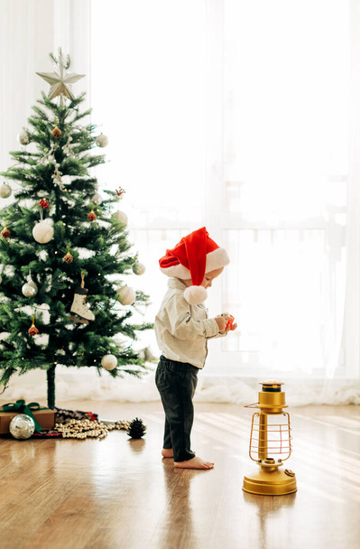 Egy kisgyerek egy karácsonyi kalapban egy karácsonyfa hátterében. Karácsonyi témájú kép, képeslap. - Fotó, kép