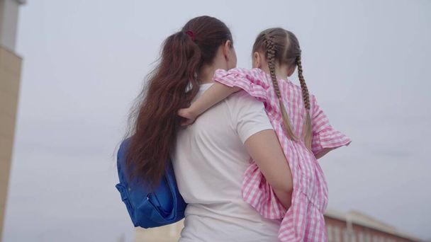 Matka niesie małą córeczkę z plecakiem w ramionach do szkoły, ucząc dziecko w podstawówce, szczęśliwą rodzinę, odprowadzając dziecko z torbą szkolną, zdobywając wiedzę o przyszłym życiu - Zdjęcie, obraz