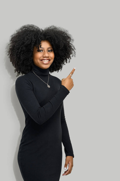 Porträt eines dunkelhäutigen Mädchens mit Afrofrisur, zeigt mit den Vorderfingern, zeigt Leerraum für ihre Promotion, isoliert auf grauem Hintergrund Menschen, Werbekonzept - Foto, Bild