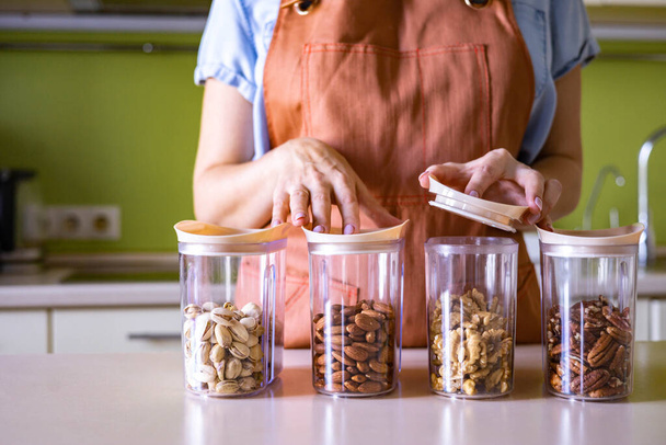 Closeup woman hands in apron placing different nuts into glass jar storage container at kitchen - Foto, Imagem