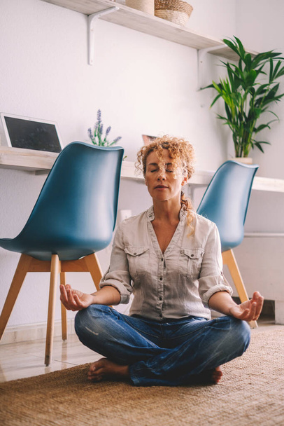 Relax en meditatie indoor activiteit volwassen jonge vrouw zitten in loto positie thuis in de woonkamer met computer werkplek op de achtergrond.  - Foto, afbeelding