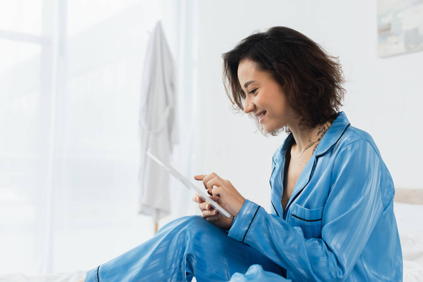 Evinde dijital tablet kullanan mavi pijamalı mutlu genç bir kadın.  - Fotoğraf, Görsel