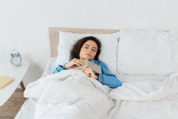 brunette vrouw in blauw pyjama slapen met boek in bed - Foto, afbeelding
