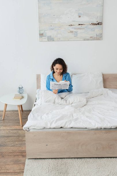alegre joven mujer en pijama leyendo viaje vida periódico y sosteniendo taza en cama - Foto, Imagen