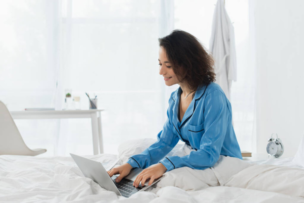 cheerful young freelancer in pajamas using laptop on bed - Foto, afbeelding