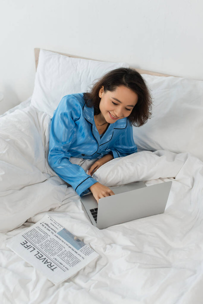 χαμογελαστός νεαρός ελεύθερος επαγγελματίας με πιτζάμες χρησιμοποιώντας φορητό υπολογιστή κοντά σε εφημερίδα στο κρεβάτι - Φωτογραφία, εικόνα