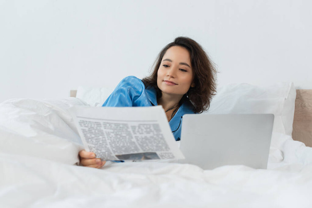 μελαχρινή νεαρή γυναίκα με πιτζάμες διαβάζει εφημερίδα κοντά σε φορητό υπολογιστή στο κρεβάτι - Φωτογραφία, εικόνα