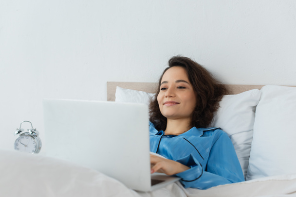 cheerful young woman in pajamas using laptop near alarm clock in bedroom - Photo, Image