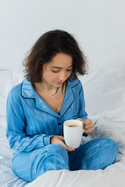 young woman in blue pajamas holding cup of coffee in bedroom  - Fotografie, Obrázek