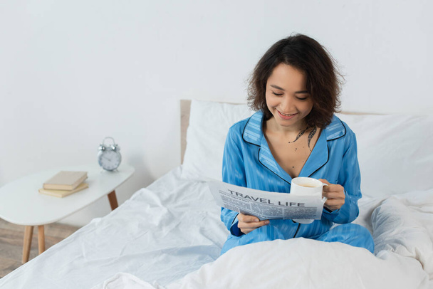 cheerful young woman in pajamas reading travel life newspaper and holding cup in bed - Фото, изображение
