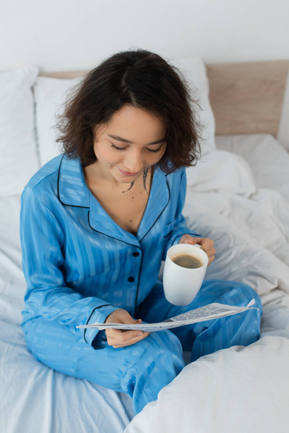 korkea kulma näkymä nuori nainen sininen pyjama tilalla kuppi kahvia lukiessaan sanomalehti makuuhuoneessa  - Valokuva, kuva