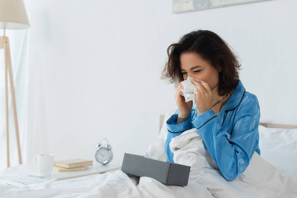 sick woman sneezing in napkin near tissue box on bed - Foto, Imagem