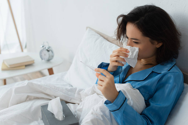 больная женщина чихает в тканях и смотрит на электронный термометр - Фото, изображение