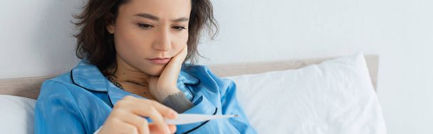 больная молодая женщина смотрит на электронный термометр, баннер - Фото, изображение