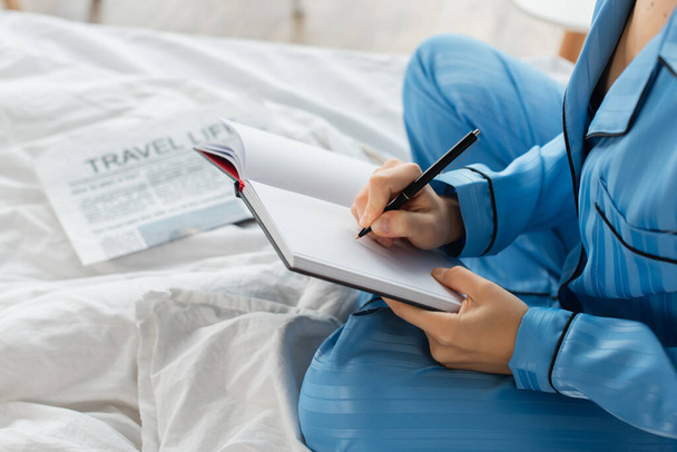 περικοπή άποψη της νεαρής γυναίκας που γράφει στο σημειωματάριο κοντά στην εφημερίδα στο κρεβάτι - Φωτογραφία, εικόνα
